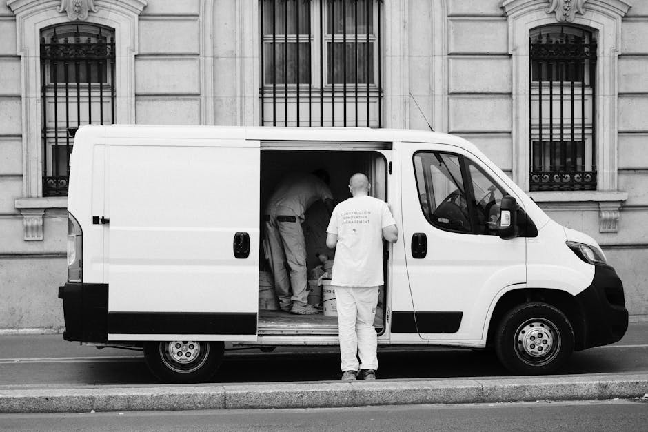 A white van parked on a city street during a home relocation process, with its rear doors open revealing a variety of packed cardboard boxes, some sealed with tape, and large plastic bags filled with 