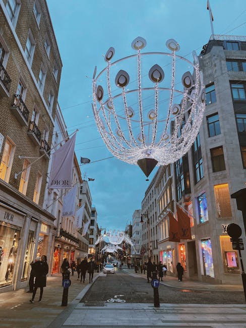 A wide street scene during daytime with a clear blue sky overhead, featuring modern and historic shopfront buildings on both sides. The storefronts display luxury brands such as Dior, with large glass