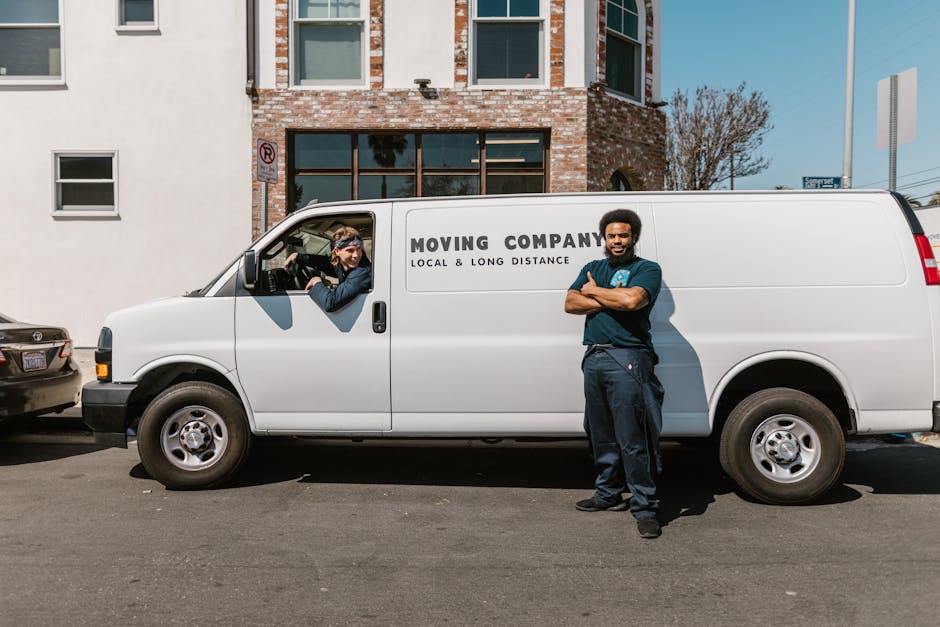 A professional house removals worker standing in front of a white moving van with the company logo 'MOVING COMPANY' and the tagline 'Local & Long Distance', parked on a street with a brick and stucco